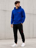 Man in een blauwe hoodie met 'PARIS' tekst en rode/zwarte streepjes, en een zwarte joggingbroek met 'PARIS' op de zijkant, staand buiten.