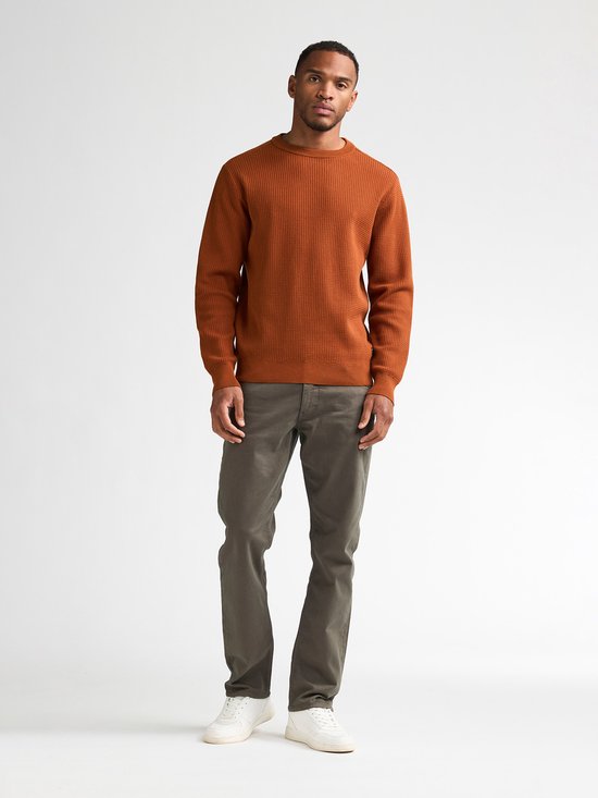 Man staat frontaal, draagt een oranje trui met ronde hals en wafelpatroon en een grijze broek.