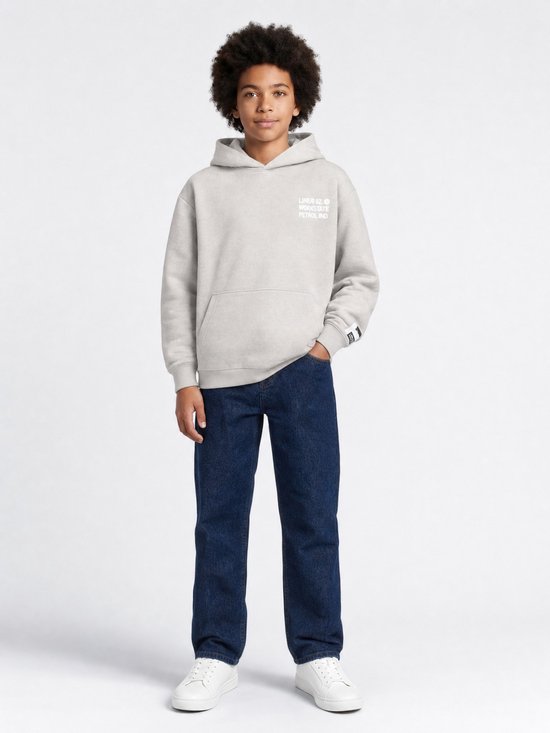 Jongen staat naar voren gericht in een lichtgrijze hoodie met witte tekst op de borst en donkere jeans. Handen in de zakken.