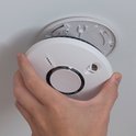Hand monteert witte rookmelder met een ronde zilveren knop op een plafondbevestiging.