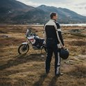 Man met helm in DXR motorjas en -broek, zwart/beige, staand naast motor in landschap.