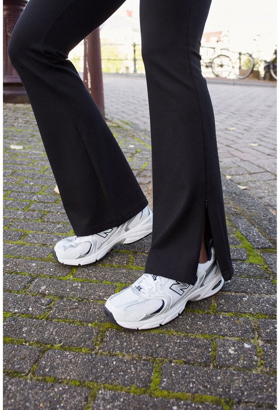 Sluiting van de split aan de onderkant van de zwarte, wijduitlopende legging, met witte sneakers.