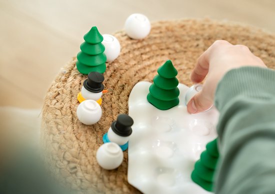 Hand legt speelstukken van "Snow Problem": sneeuwpoppen, bomen en witte ballen op spelbord.