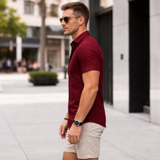 Man draagt een bordeauxrood overhemd met korte mouwen en beige short. Hij draagt zonnebril en horloge.