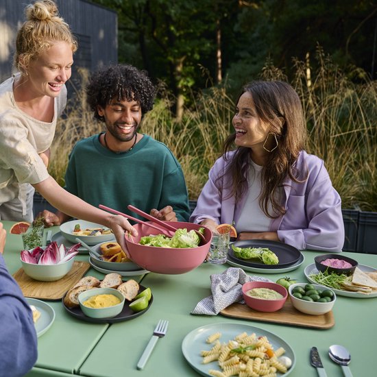 Een vrouw serveert salade uit een roze kom aan tafel terwijl vrienden lachen.
