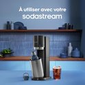 Sodastream bruiswatermaker met een blauwe Sodastream fles erin. Rechts een glas bruisende cola met ijs en limoen.