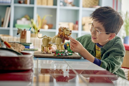 Een jongen met een bril bouwt een LEGO leeuw op een tafel. De leeuw is gemaakt van bruine en beige stenen.
