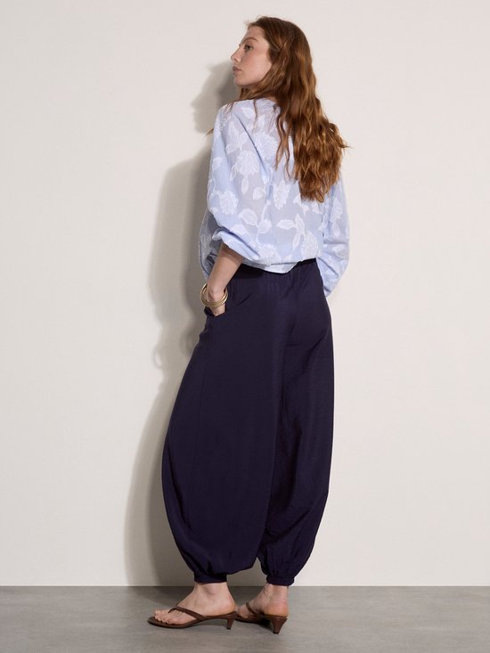 Model van achteren gezien in losvallende blauw gestreepte blouse met bloemenpatroon en donkere ballonbroek.