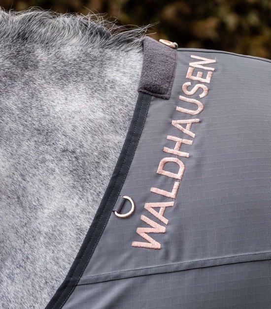 Close-up van een grijze paardendeken met de tekst "WALDHAUSEN" in koperkleurige letters.