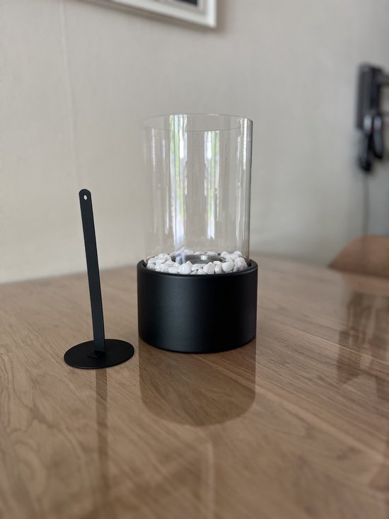 Zwarte tafelhaard met glazen cilinder, witte steentjes en een zwarte staander op een houten tafel.