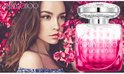Vrouw naast een fles Jimmy Choo Blossom parfum tegen een achtergrond van roze bloemen.