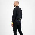 Man met baard, staand in profiel, draagt zwart fietsjack met fluor accenten en zwarte fietsbroek.