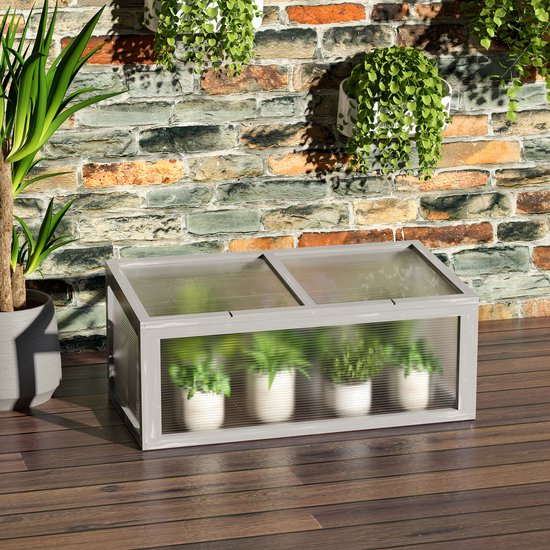 Grijze houten mini-kas met planten in witte potten, geplaatst op een houten terras tegen een bakstenen muur.
