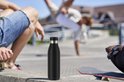 Zwarte Tefal Bludrop thermosfles met zilverkleurige ring, in de voorgrond op een betonnen rand, met skaters op de achtergrond.