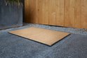 Kokosdeurmat met zwarte rand op een betonnen ondergrond, naast een houten deur.