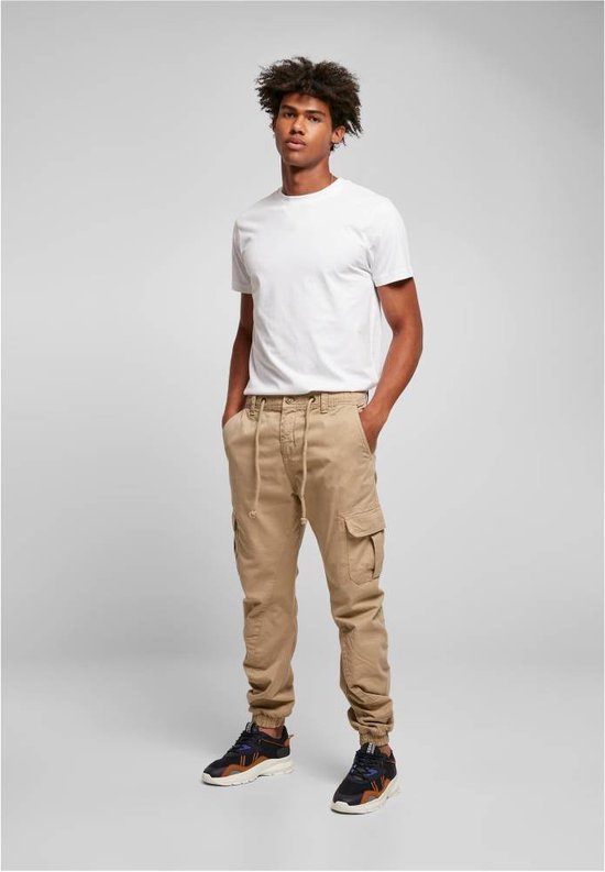 Man staat in beige cargo broek, met trekkoord, zijzakken en elastische enkelbanden.