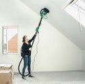 Vrouw schuurt een schuin plafond met een lange groene schuurmachine.