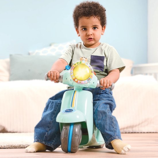 Zittende jongen met spijkerbroek en lichtgroene driewielige scooter met blauw zadel.