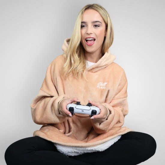 Een vrouw met blond haar zit met gekruiste benen en speelt een videogame. Ze draagt een oversized beige hoodie deken.