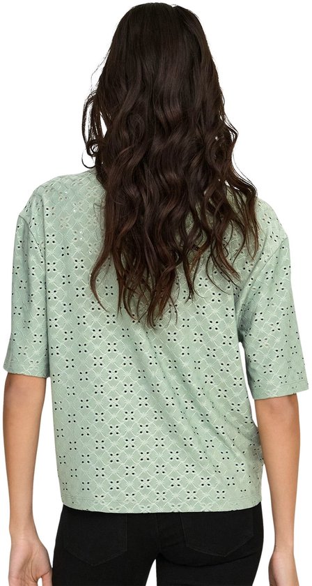 Achterkant van een vrouw in een lichtgroene blouse met broderie anglaise en driekwart mouwen, met zwarte broek.
