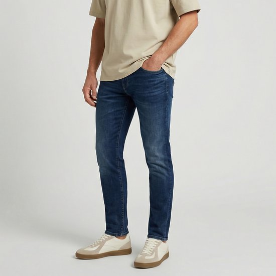 Mannelijke benen in een donkerblauwe, slank gesneden spijkerbroek en beige sneakers, met de hand in de zak.