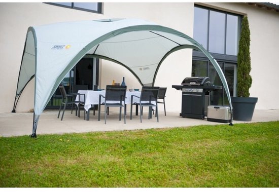 Een Coleman partytent met tafel en stoelen, naast een huis met een barbecue.