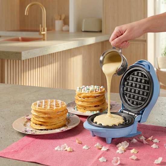 Blauw wafelijzer met beslag dat wordt gegoten. Wafels gestapeld op een roze schaal met bloemenpatroon.