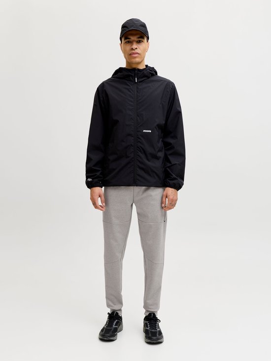 Man met zwarte jas, grijze joggingbroek, pet en sneakers. Frontaal zicht op witte achtergrond.