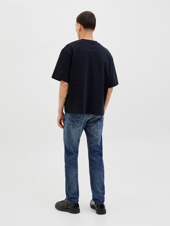 Man in een zwarte oversized T-shirt en blauwe jeans, met de rug naar de camera.