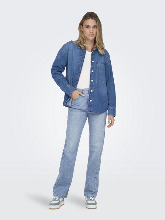 Vrouw in een witte tanktop, lichtblauwe straight leg jeans en een denim blouse, staand.