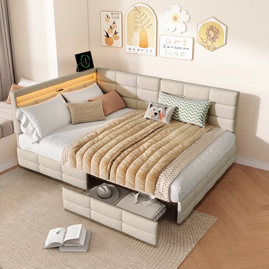 L-vormig beige linnen bed met gestoffeerde hoofdborden, opbergruimte, dekens, kussens en nachtkastje met lamp.