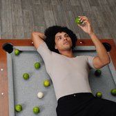 Man met afro kapsel ligt op een pooltafel, draagt een beige v-hals ondershirt en houdt een limoen vast.