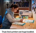 Vrouw met blauwe hoodie zit aan een houten bureau en gebruikt een HP printer om thuis hoge kwaliteit foto's te printen.