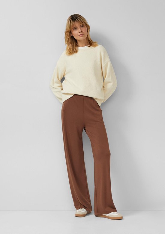 Model draagt een bruine wijde broek en een gebreide crème trui, poserend met handen achter de rug.