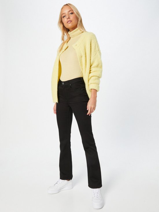 Vrouw in zwarte flared jeans, lichtgele coltrui en geel vest, met witte sneakers.