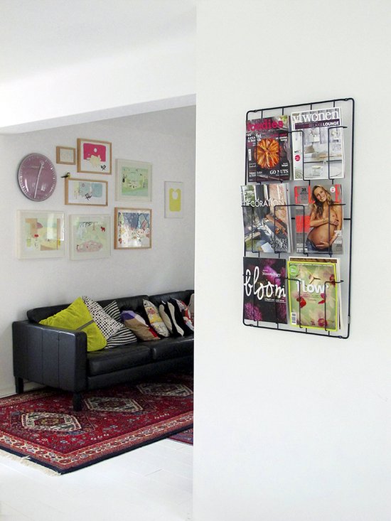 Een zwart tijdschriftenrek met 6 vakken, gevuld met tijdschriften, hangt aan de muur naast een bank.