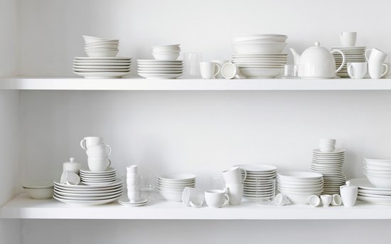 Wit porseleinen serviesgoed, waaronder stapels borden, schalen, mokken en theepotten, op witte planken.