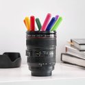Zwarte cameralens beker gevuld met gekleurde stiften, op een bureau naast boeken.