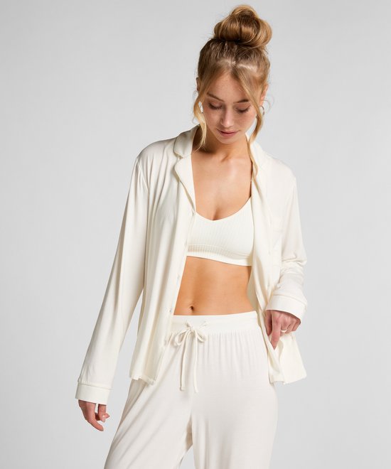 Model met knot draagt open wit pyjamashirt over een witte top en broek.