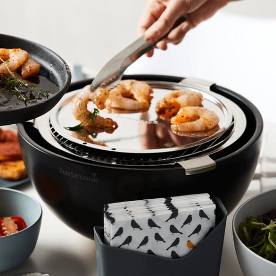 Zwarte tafelgrill met gegrilde garnalen, een hand legt er met een tang nog meer op.