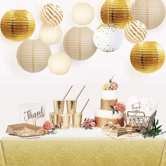 Een feesttafel met een gouden glitter tafelkleed, gouden en witte lampionnen, gouden bekers, cadeautjes en een bruidstaart.