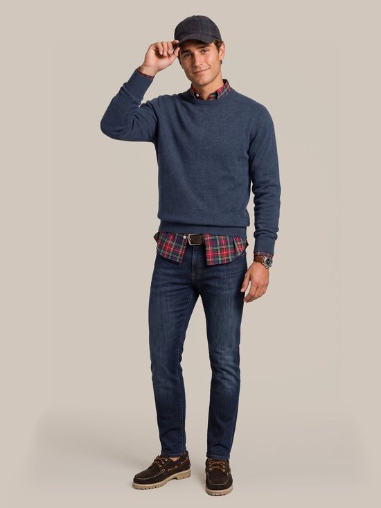 Model draagt een blauwe lamswollen trui over een rood geruit overhemd, donkere jeans, bootschoenen en een pet.