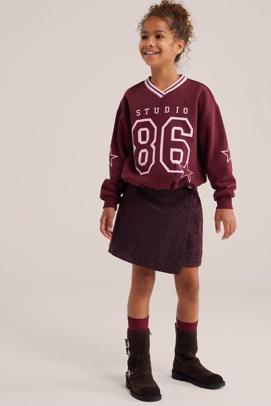 Meisje met krullend haar in een bordeauxrode sweater met 'STUDIO 86' en een bordeauxrode glitter wikkelrok.