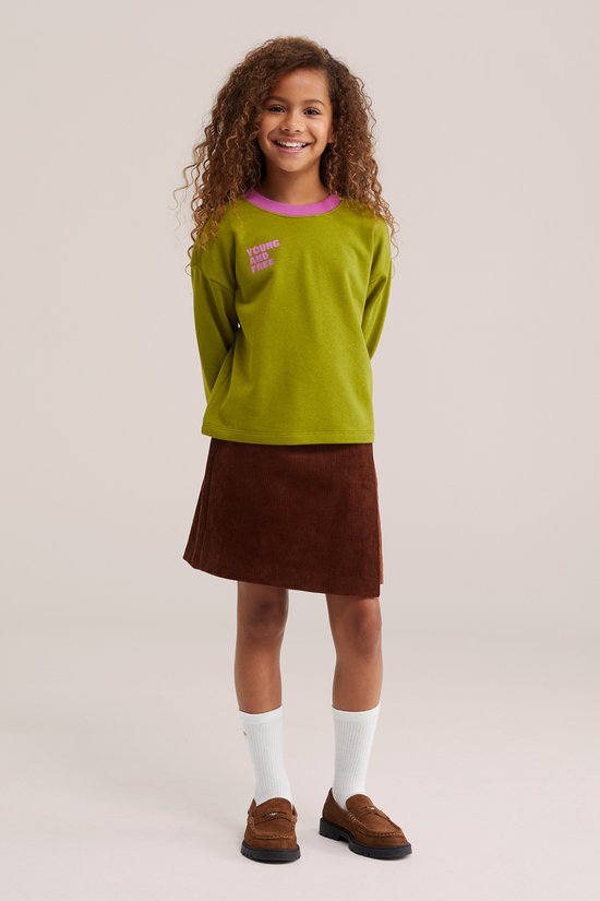 Lachend meisje in groen shirt met tekst, bruine corduroy rok met overslag, witte sokken en bruine mocassins.