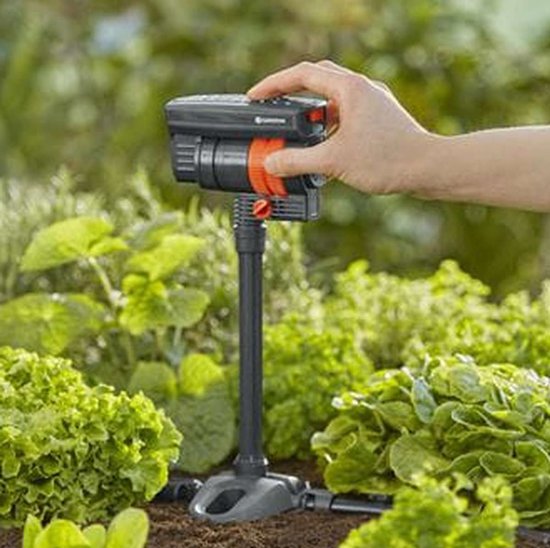 Een hand past de grijze GARDENA zwenksproeier met oranje accenten aan in een groentetuin.