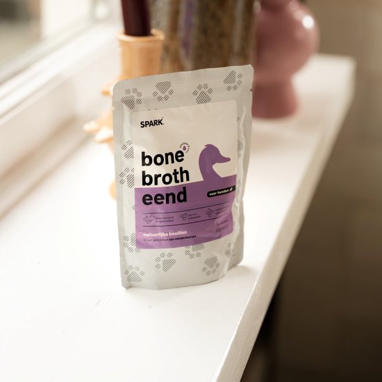 Pakje 'bone broth eend' met paarse eend, op een witte vensterbank met decoraties.