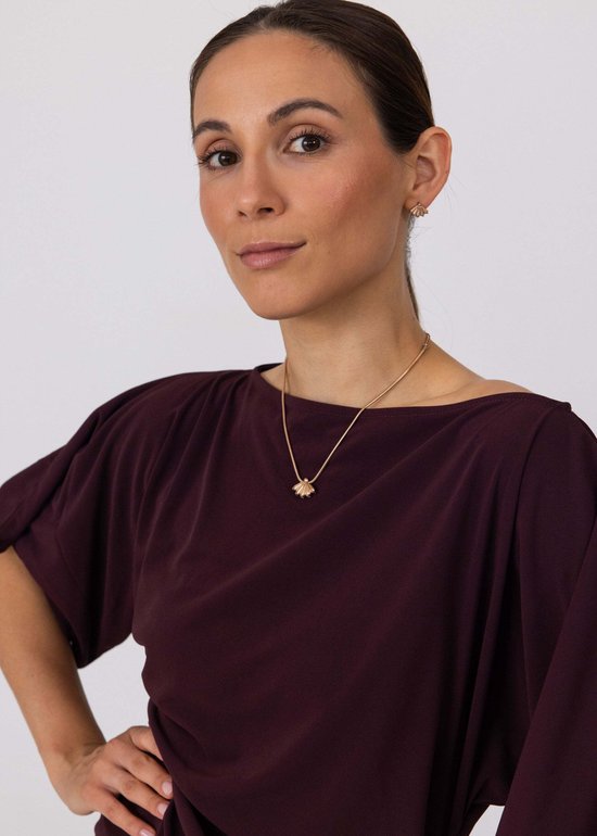 Model met gouden Purelei ketting met bloemhanger en bijpassende oorbellen. Draagt een elegante paarse top.