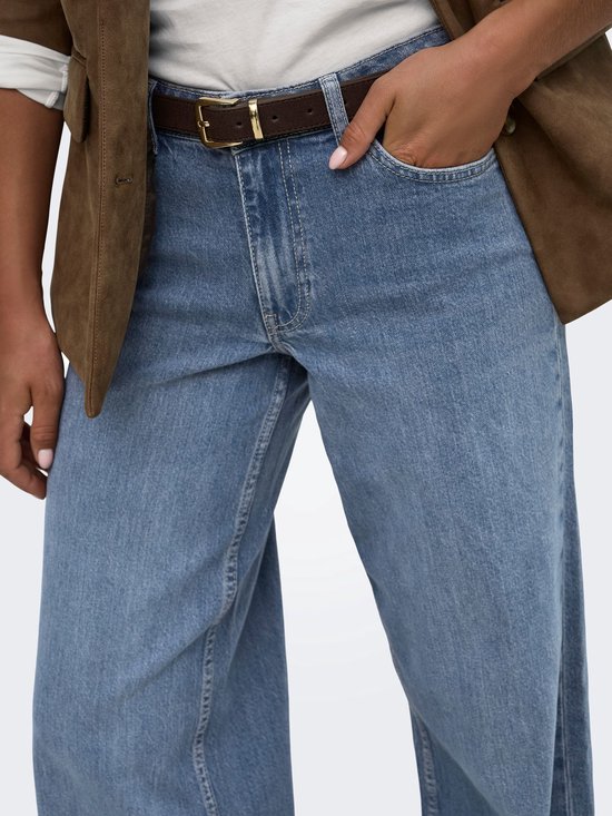 Detailopname van de tailleband en zakken van een spijkerbroek met wijde pijpen, gedragen met een witte top en bruine blazer.