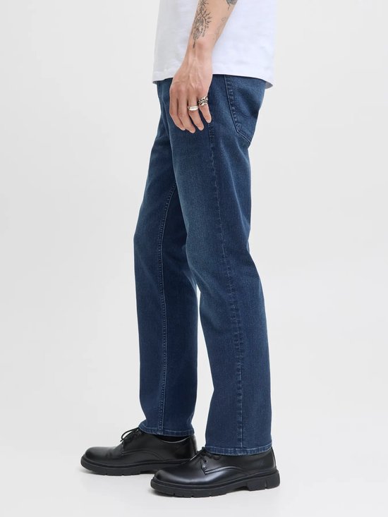 Zijaanzicht van een man met tatoeages in een witte T-shirt en een donkerblauwe jeans.