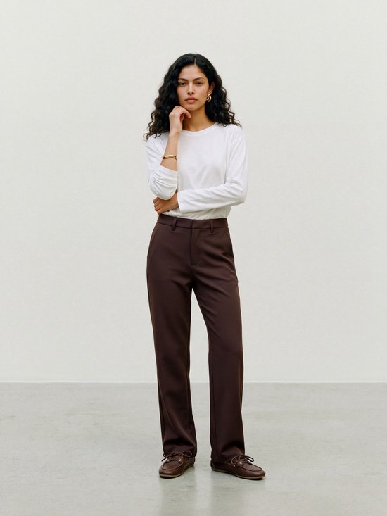 Model in een wit shirt met lange mouwen, donkerbruine pantalon en bruine schoenen, hand onder de kin.